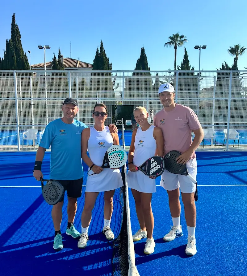 Vier spelers met padelrackets bij het net op een blauwe padelbaan