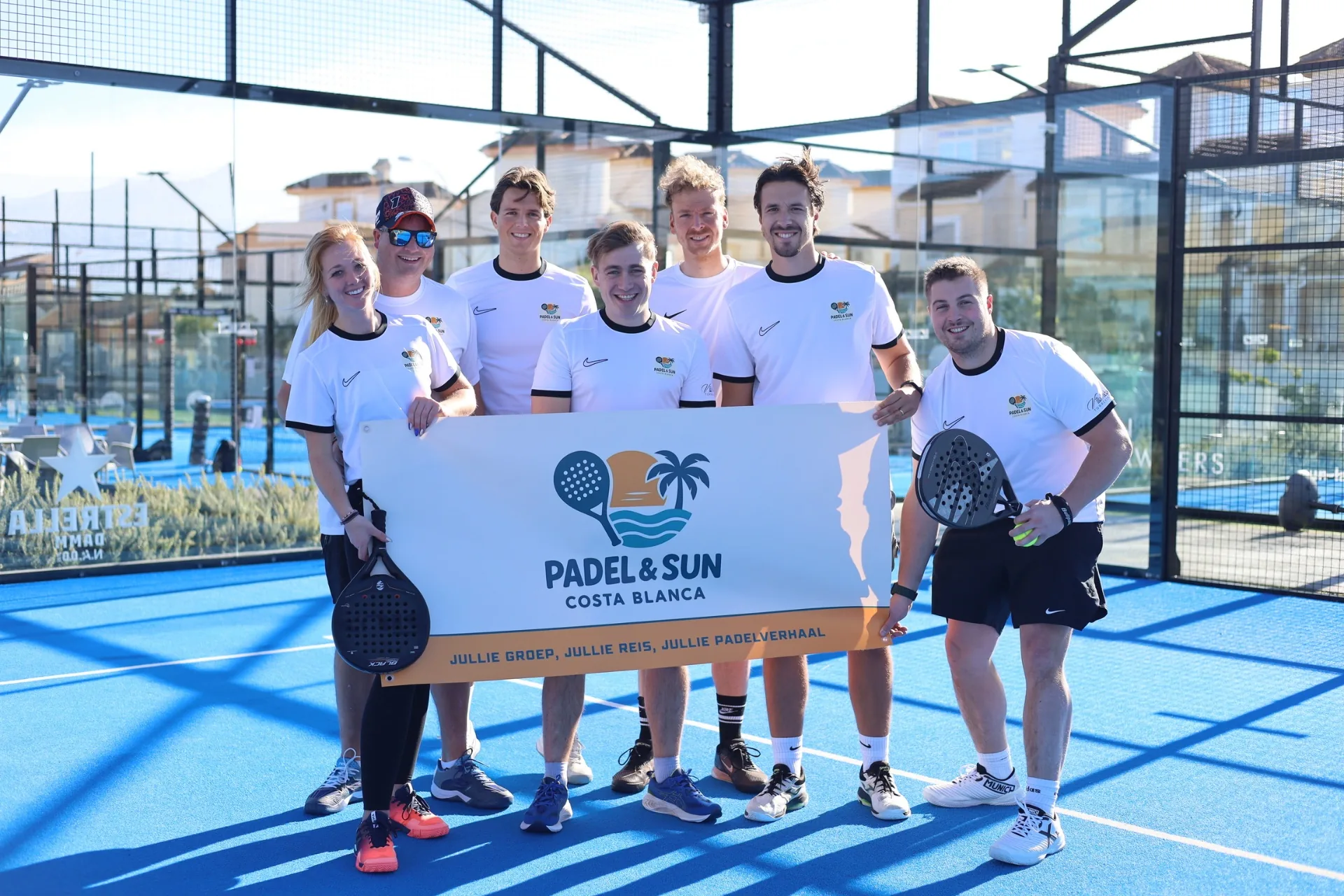 Groep deelnemers poseert samen op de padelbaan met Padel & Sun bord